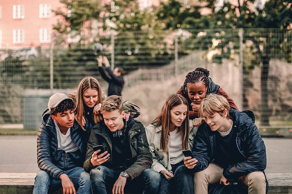 Teenager-Gruppe sitzt draußen auf einer Bank und schaut auf zwei Smartphones.