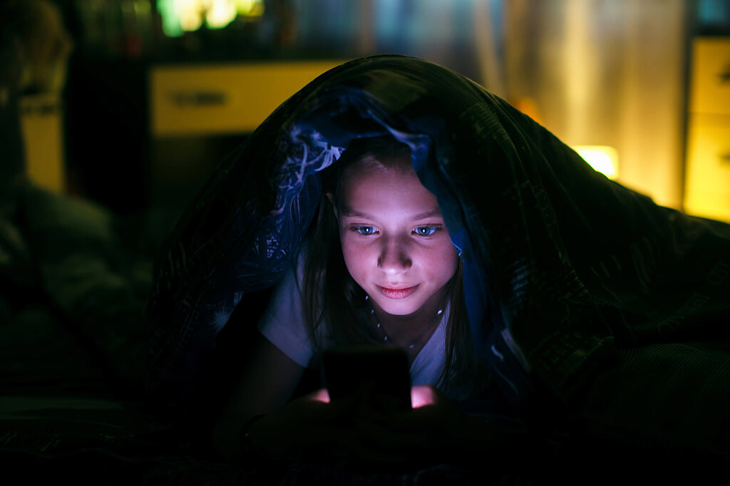 Mädchen schaut im Dunkeln unter der Decke auf ihr Smartphone. 