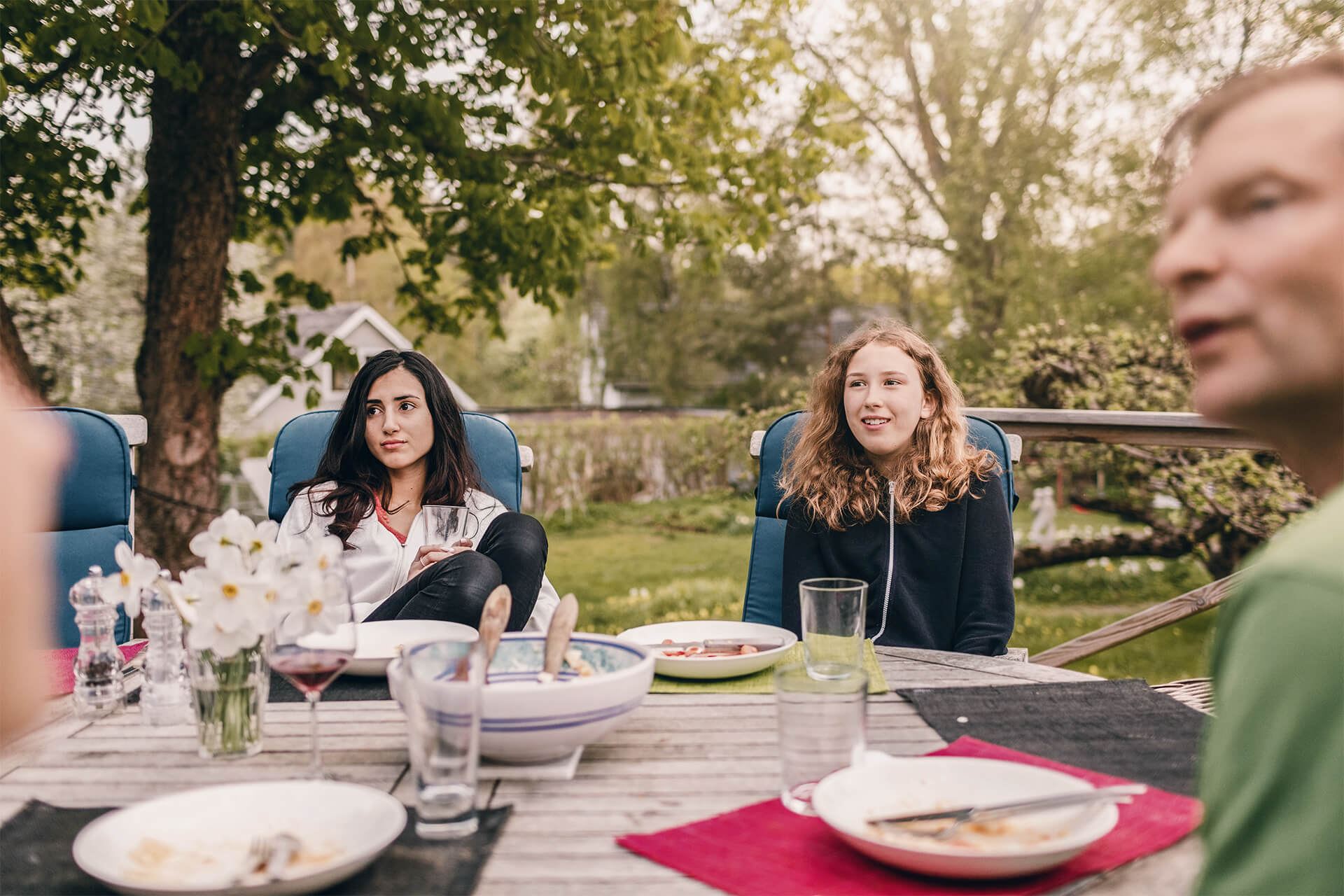 Zwei Teenagerinnen sitzen mit einem Erwachsenen an einem Tisch im Garten.
