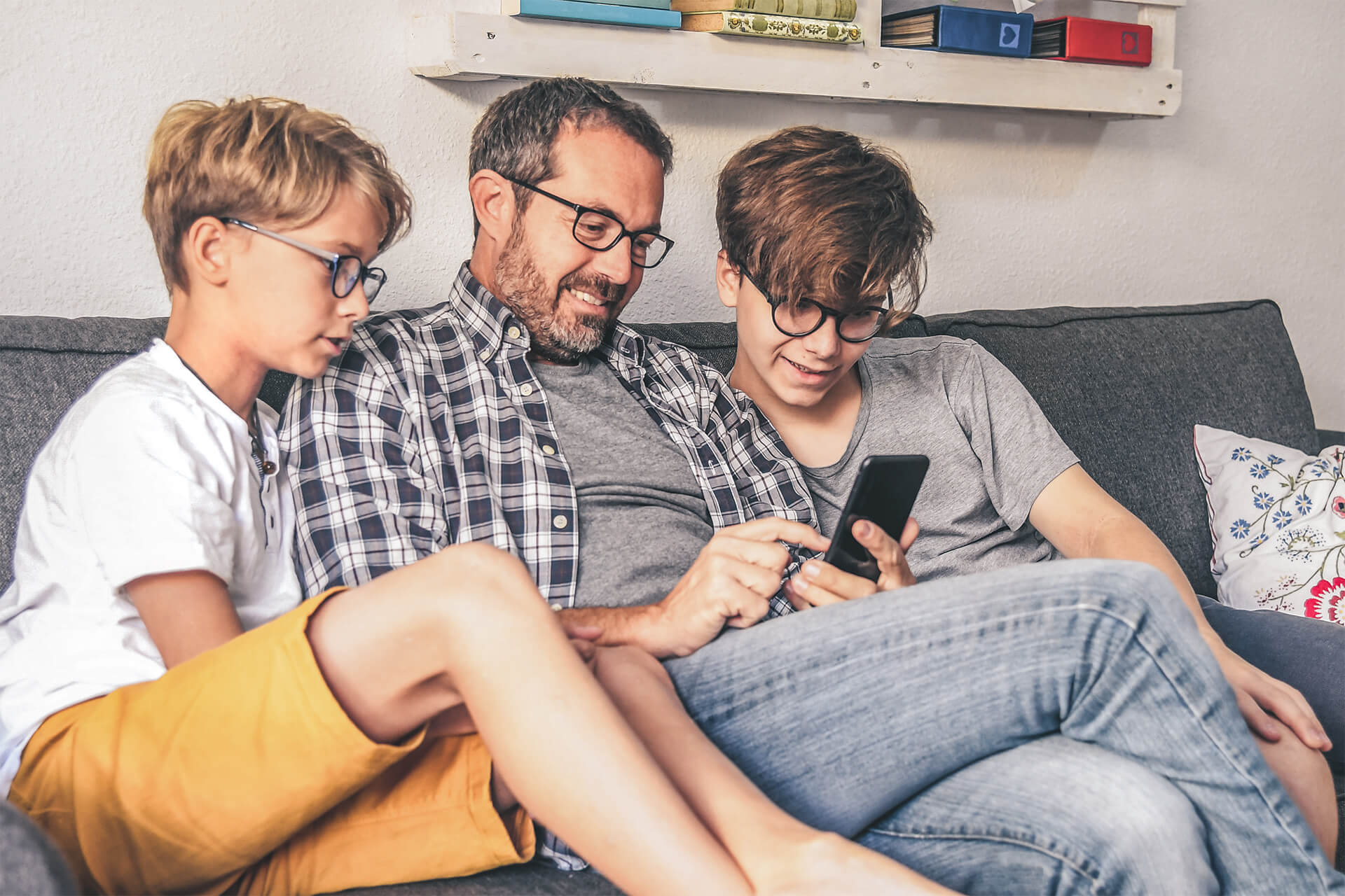 Vater sitzt mit seinen zwei Söhnen auf der Couch und zeigt etwas auf seinem Handy.